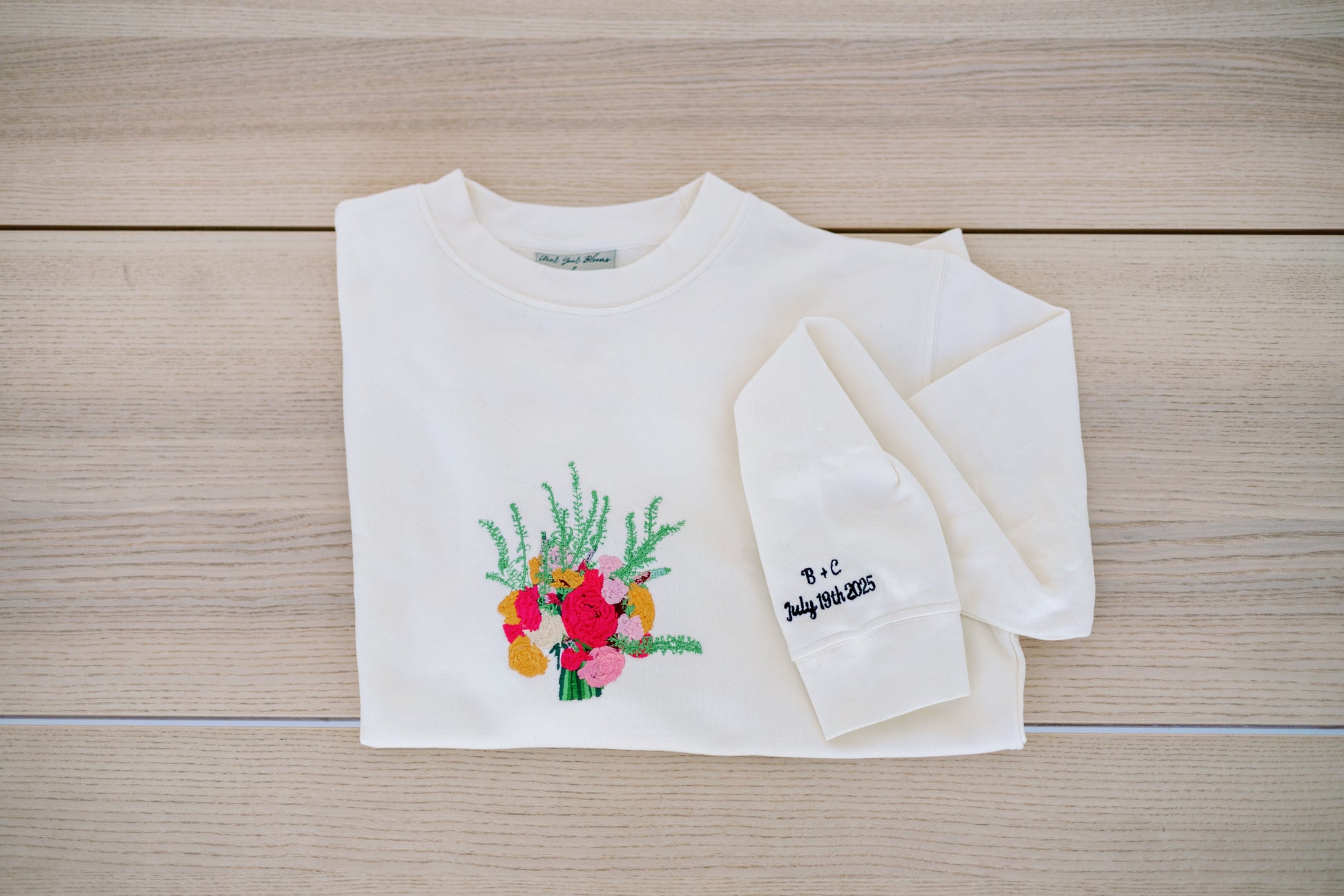 Custom embroidered bouquet crewneck in ivory color laid flat with name and date on sleeve