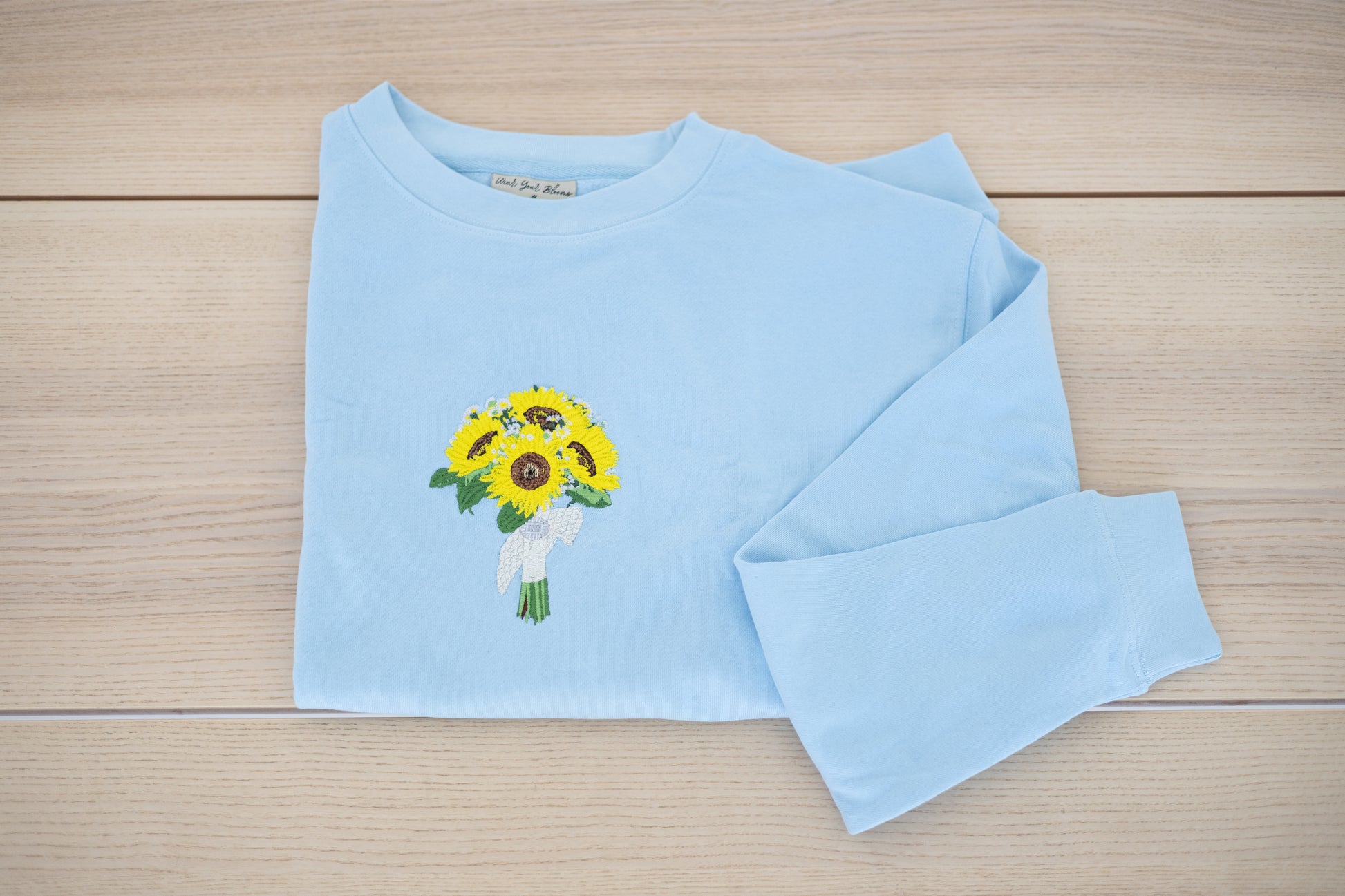 Embroidered wedding bouquet crewneck in light blue laid flat showing sunflower design